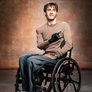A young man with a prosthetic hand sits in a wheelchair, smiling confidently. He is wearing a beige long-sleeve shirt and jeans, with a warm, blurred background.