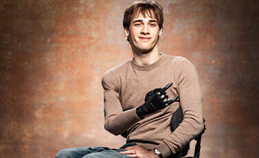 A young man with a prosthetic hand sits in a wheelchair, smiling at the camera. He wears a tan long-sleeve shirt and jeans, with a neutral studio backdrop behind him.
