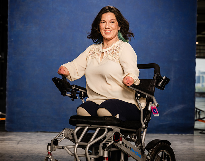 A smiling woman with short dark hair, wearing a cream blouse, sits in a motorized wheelchair. She is a quadruple amputee, and the background is a plain blue backdrop.