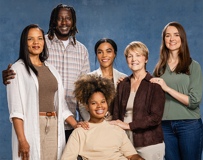 A diverse group of six people, including men and women of different ages and backgrounds, stand and sit smiling together against a blue textured background. One young person sits in front while the others stand behind.
