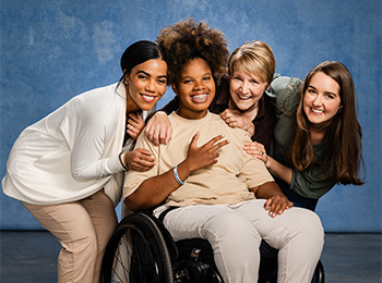 Four women smile together in front of a blue background; one is seated in a wheelchair while the other three crouch beside her, showing support and friendship.