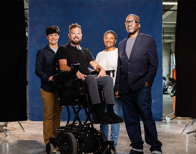 Four people pose together in front of a blue backdrop. One person is seated in a power wheelchair, while the others stand close by, smiling. The setting appears to be an indoor studio with equipment visible in the background.