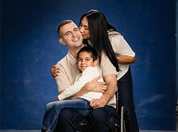 A man in a wheelchair hugs a young boy on his lap while a woman stands behind, kissing his cheek. All three are smiling against a blue background.
