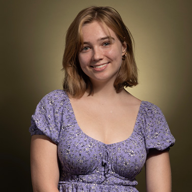 A young woman with light brown hair, wearing a short-sleeve purple floral dress, smiles and poses in front of a brownish-yellow gradient background.