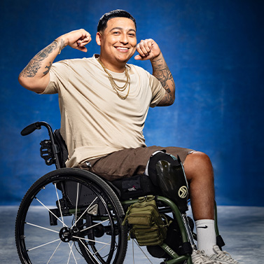 A smiling man with short dark hair sits in a wheelchair, flexing his arms. He wears a beige t-shirt, shorts, white socks, and gold chains, with a blue background behind him.