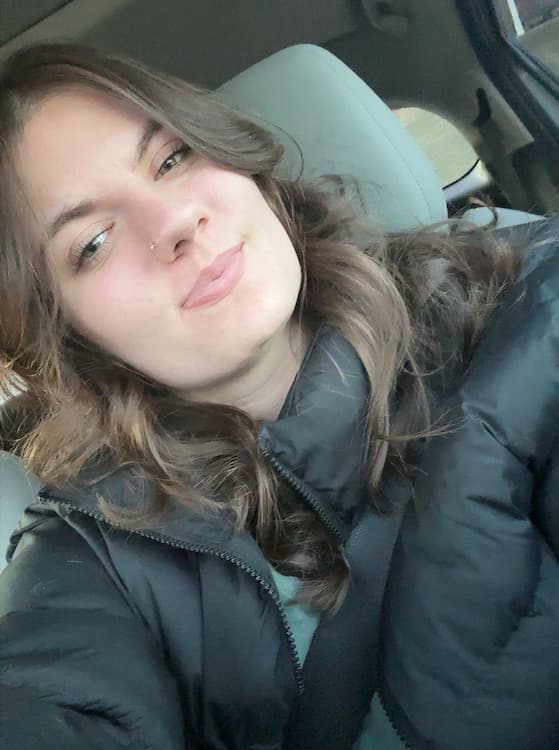 A woman with wavy brown hair and a nose piercing takes a selfie in a car. She is wearing a black puffer jacket and is seated with a seatbelt on, smiling softly at the camera.