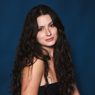 A young woman with long, wavy brown hair poses in front of a dark blue background, wearing a sleeveless black top and softly smiling at the camera.