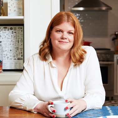 A woman with shoulder-length red hair and a white blouse sits at a table holding a mug, smiling. The background shows a kitchen and shelves with books or decorative items.