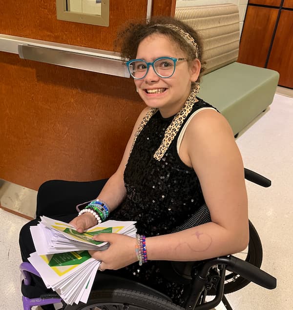 A smiling young girl with glasses, wearing a black sequin dress and colorful bracelets, sits in a wheelchair holding a stack of programs in her lap.