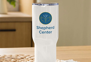 A white tumbler with a blue and yellow Shepherd Center logo sits on a wooden table.