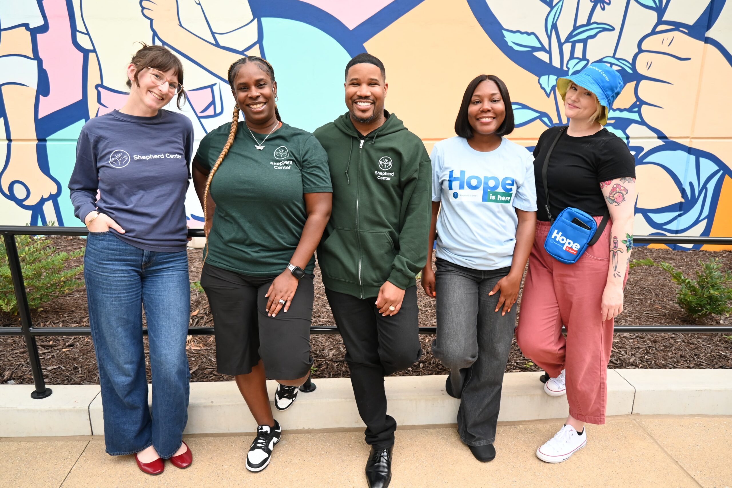 Five people stand smiling in front of a colorful mural. They wear casual clothes, including shirts and accessories with positivity messages like 
