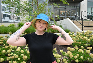 A person with tattoos on both arms smiles and holds the brim of a blue hat, standing in front of a building with stairs and green shrubs.
