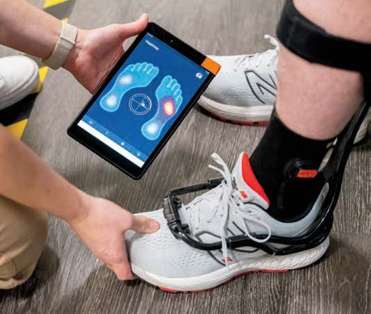 A person holds a tablet displaying a foot pressure analysis next to another person’s foot, which is wearing a white sneaker and an orthopedic brace. The screen shows highlighted pressure points on a foot diagram.
