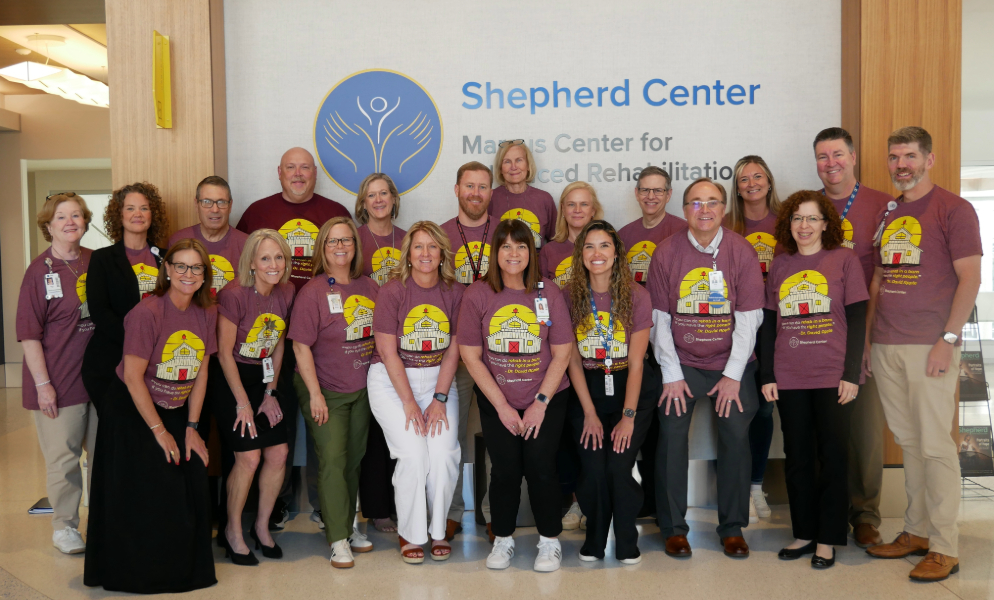 A group of 21 adults, many wearing matching maroon t-shirts with a yellow house graphic, pose and smile in front of a sign reading “Shepherd Center Marcus Center for Advanced Rehabilitation.”.