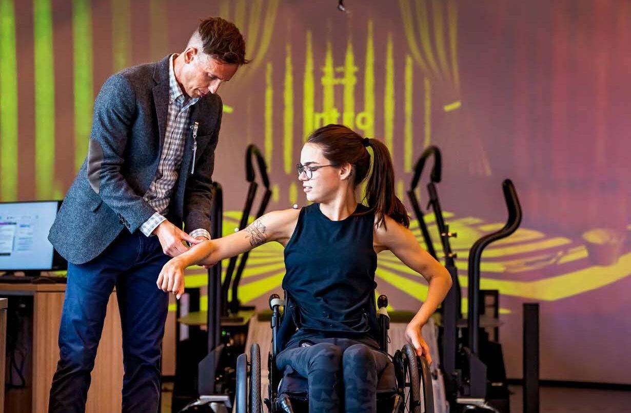 A man assists a woman in a wheelchair with arm exercises in a modern rehab facility.