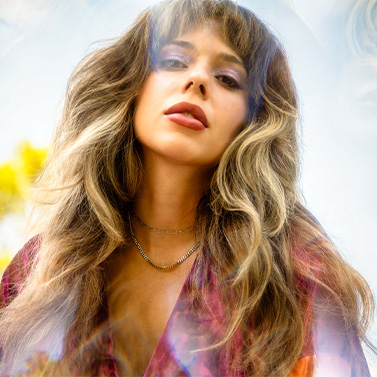 A woman with long, wavy hair and subtle highlights gazes confidently at the camera, wearing a red patterned top and layered necklaces, with a soft, dreamy light effect in the background.