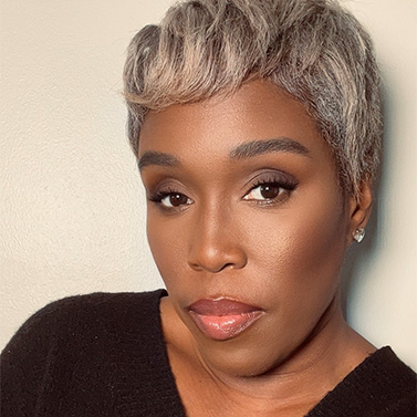 A person with short, silver and black hair, wearing subtle makeup and a black top, poses in front of a light background.