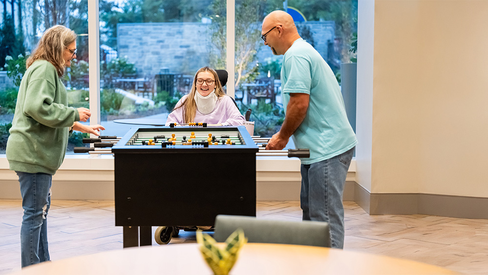 Three people play foosball indoors. A person in a wheelchair is in the middle, smiling, while two others on either side engage in the game. Large windows display a garden view in the background.
