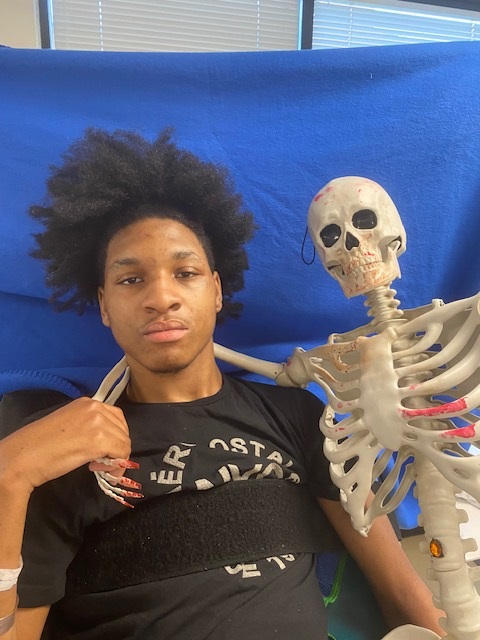 A teenage boy with sits next to a human skeleton model. The skeleton's hand rests on their shoulder, and both face the camera.